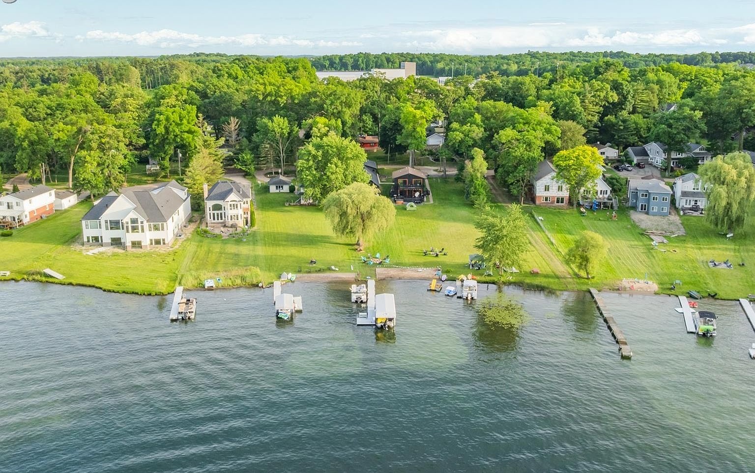 Aerial drone view of the Sugden Lake shoreline showing rolling docks, mowed lawns running down to the water, and a wooded backdrop of mature deciduous trees