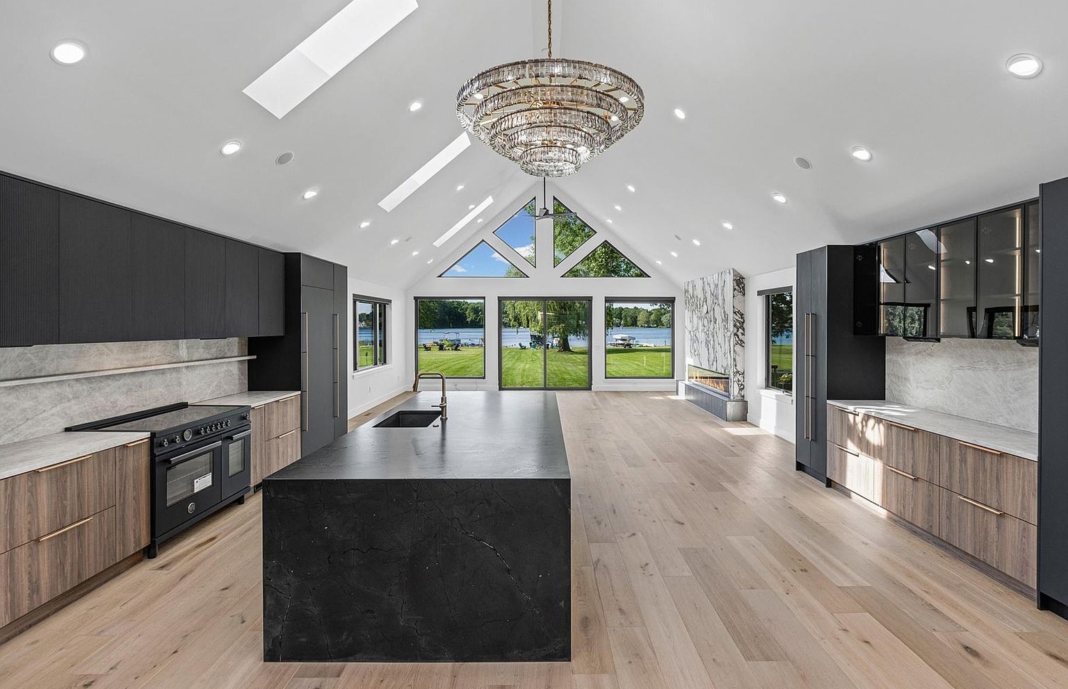 Interior of the great room: vaulted A-frame ceiling, large crystal tiered chandelier, dark slate-style waterfall island, light hardwood floors, with the triangular A-frame window wall opening onto a lawn and the lake beyond