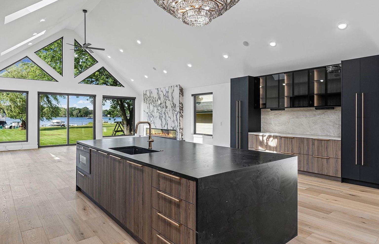 Wide angle of the great room from a different vantage: vaulted A-frame ceiling with a ceiling fan, dramatic veined marble waterfall feature, walnut and dark cabinetry, and the floor-to-peak window wall opening onto the lawn and Sugden Lake