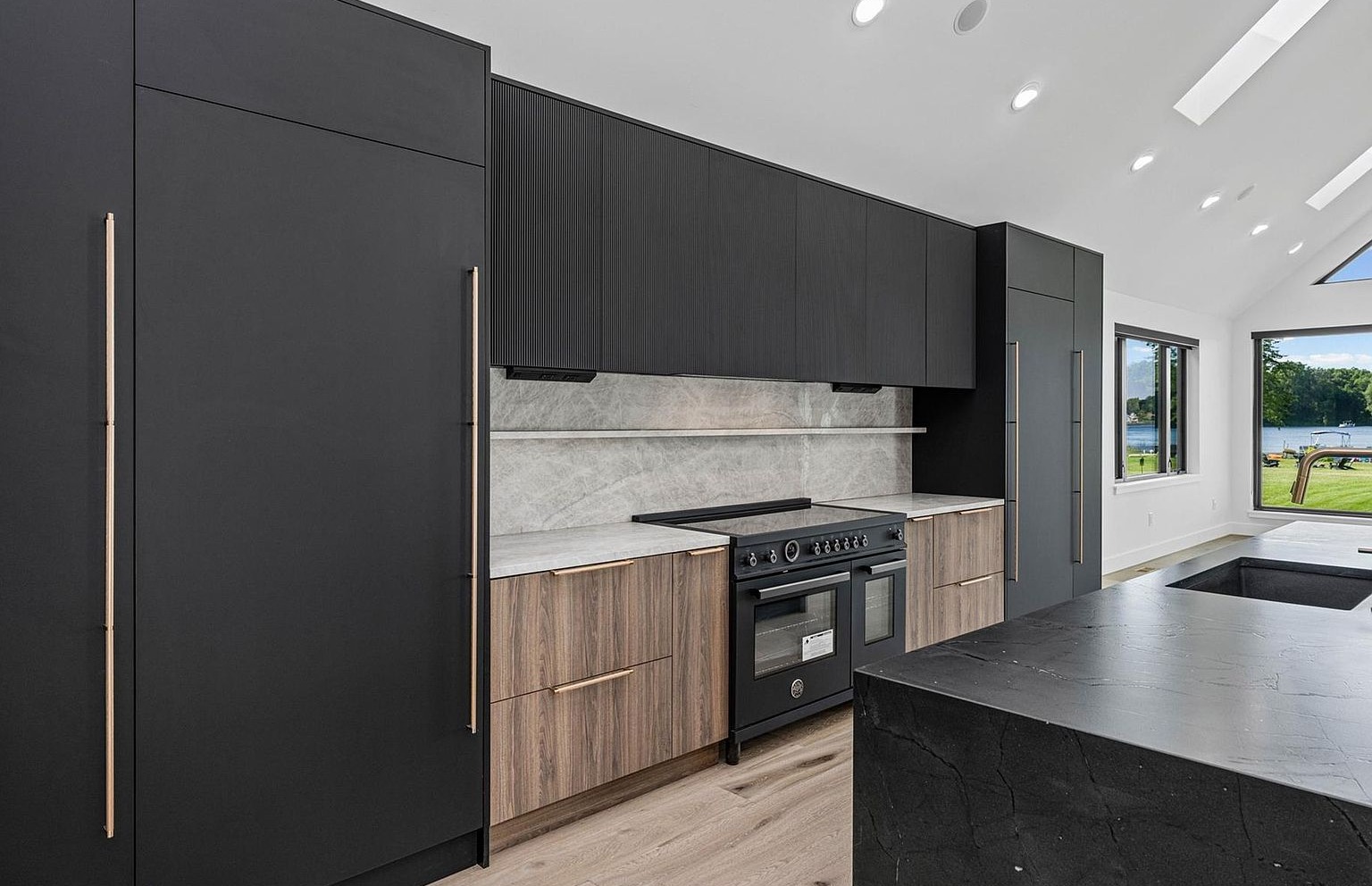 Detail of the chef's kitchen showing a black Bertazzoni induction range with red dials, dark fluted upper cabinets, light wood lower cabinets with vertical bronze pulls, a marble slab backsplash, and the corner of a dark stone island