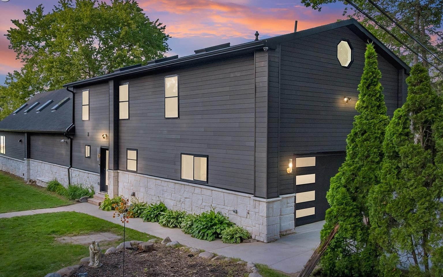 Twilight exterior of 10890 Hillway Dr, a dark charcoal vertical-siding A-frame with a stone foundation, octagonal gable window, and warm interior lights against a pink-and-orange sunset sky